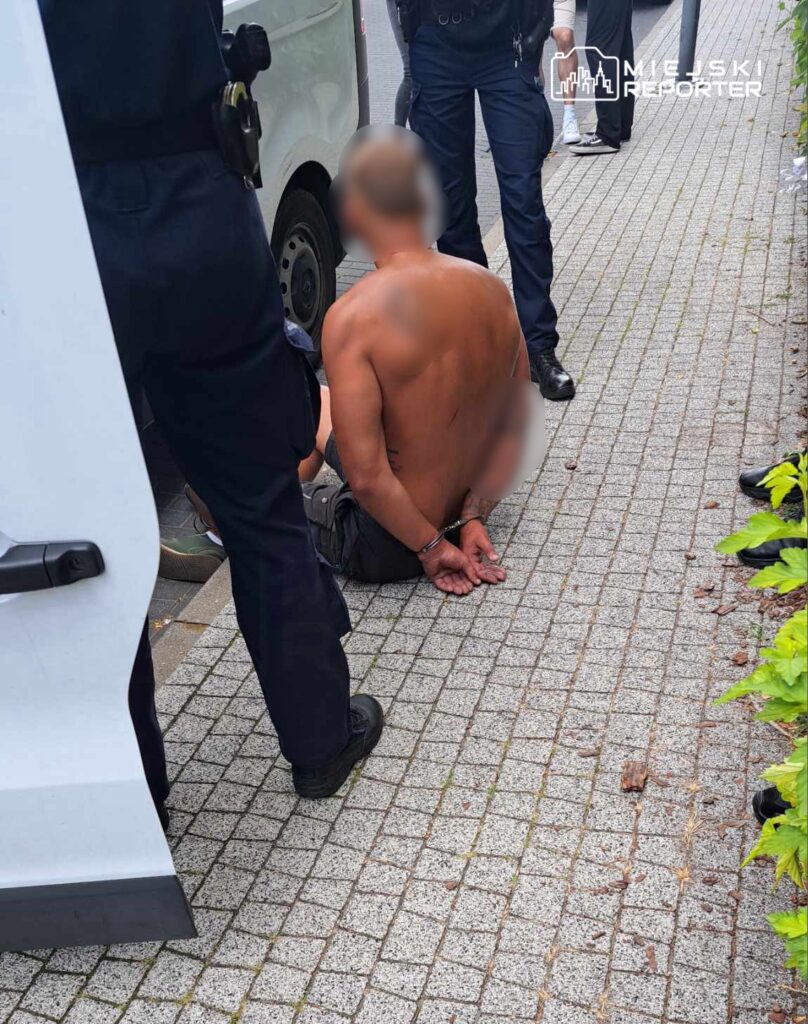 Mężczyzna w kajdankach siedzący na chodniku, otoczony przez policjantów w umundurowaniu.