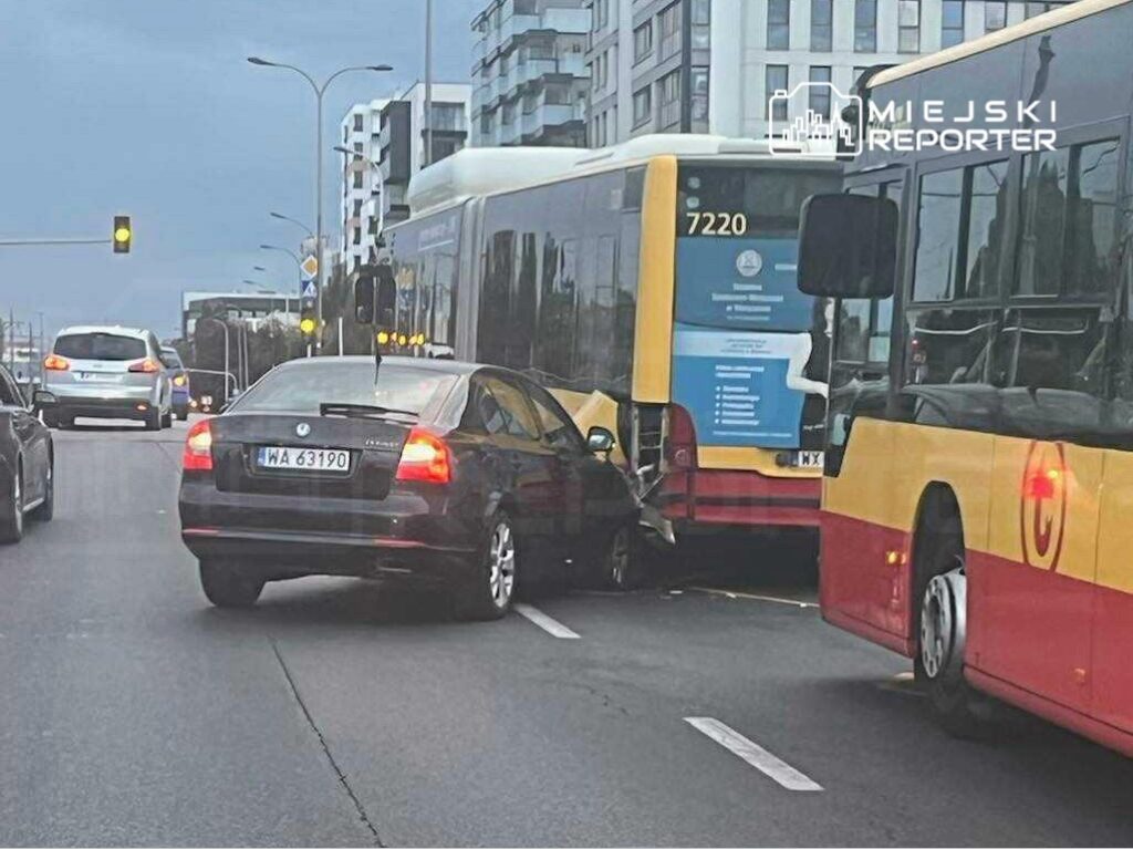 Czarny samochód osobowy uderza w tył autobusu miejskiego na ruchliwej ulicy, w tle widoczne budynki mieszkalne.