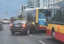 Czarny samochód osobowy uderza w tył autobusu miejskiego na ruchliwej ulicy, w tle widoczne budynki mieszkalne.