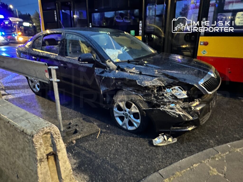 Czarny samochód osobowy z uszkodzoną przednią częścią stoi przy barierce na drodze, obok zatrzymany autobus.
