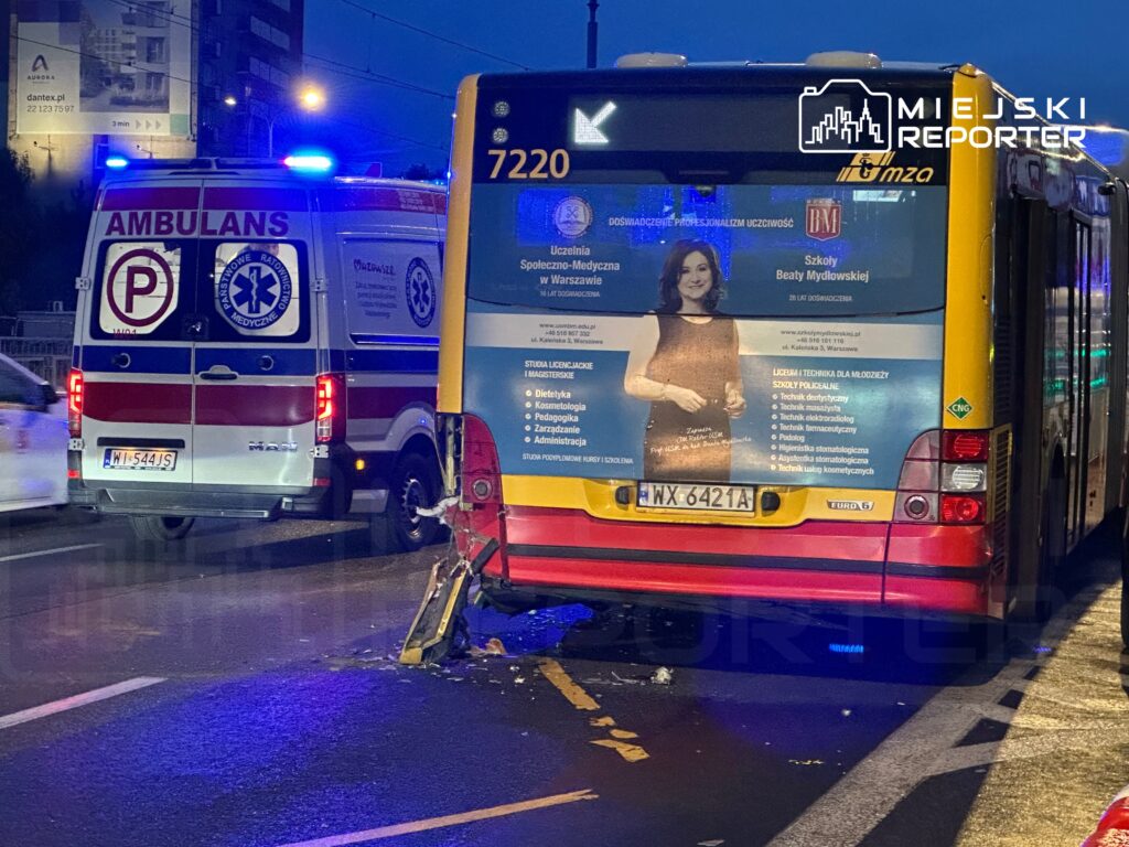 Ambulans z włączonymi sygnałami świetlnymi stoi obok uszkodzonego autobusu, na drodze w miejskim otoczeniu.