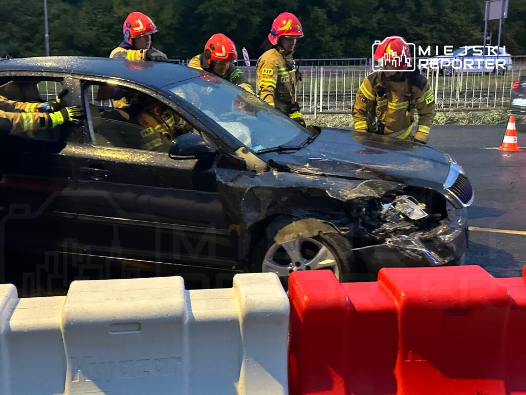 Grupa strażaków w hełmach ocenia uszkodzenia czarnego samochodu po wypadku drogowym na drodze.