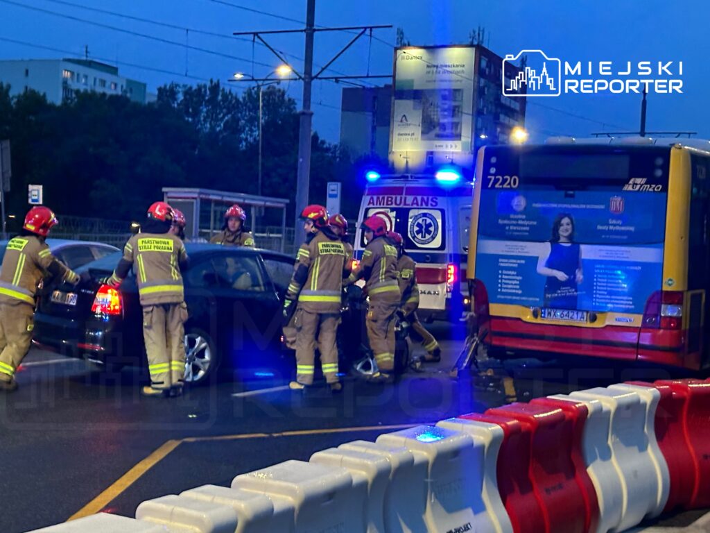 Grupa strażaków w hełmach pomaga przy wypadku drogowym, obok stoi ambulans i autobus. W tle widoczne blokady drogowe.