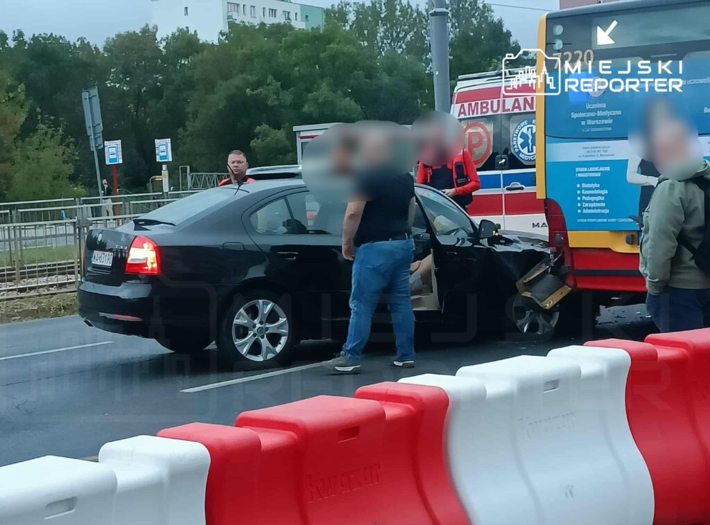 Czarny samochód osobowy z uszkodzeniami stoi przy przystanku autobusowym, a obok grupka osób rozmawia z ratownikami medycznymi.