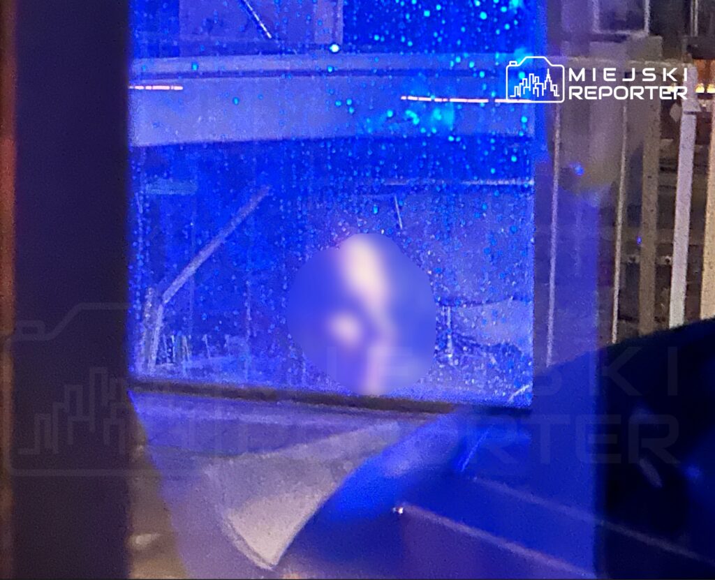 Niebieskie światło odbijające się od mokrej szyby, w tle widoczne konstrukcje budowlane i fragmenty ulicy.