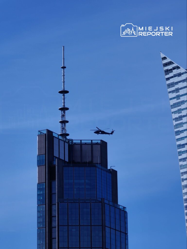Czarny helikopter przelatuje obok nowoczesnego biurowca z przeszkloną fasadą i anteną na dachu.