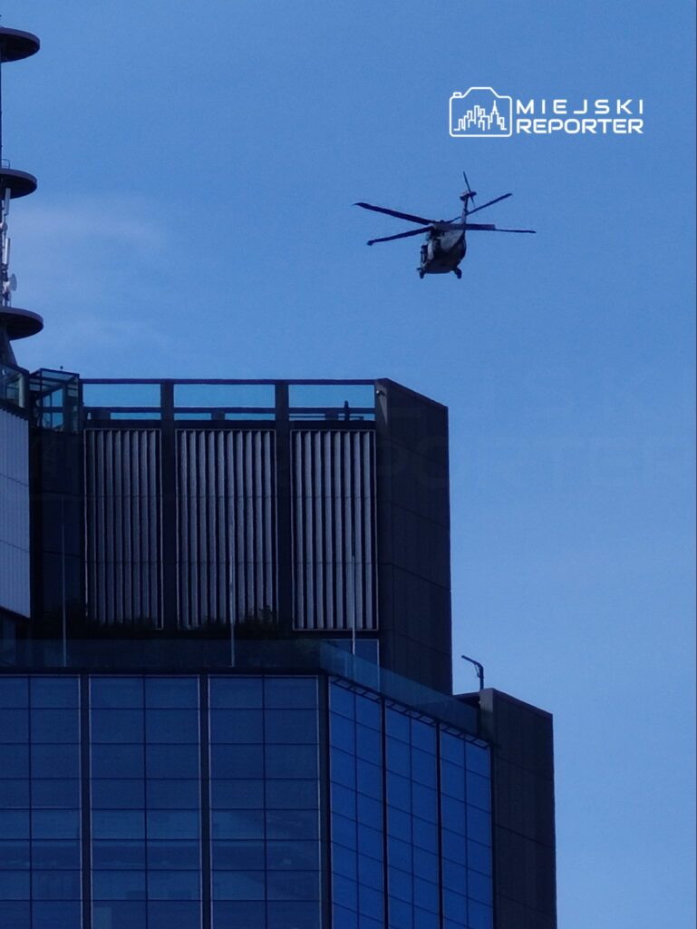 Helikopter przelatujący nad nowoczesnym biurowcem z przeszkloną fasadą w słoneczny dzień.