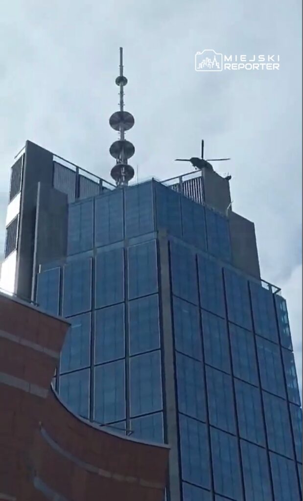 Helikopter ląduje na dachu nowoczesnego biurowca z przeszkloną fasadą w pochmurny dzień.