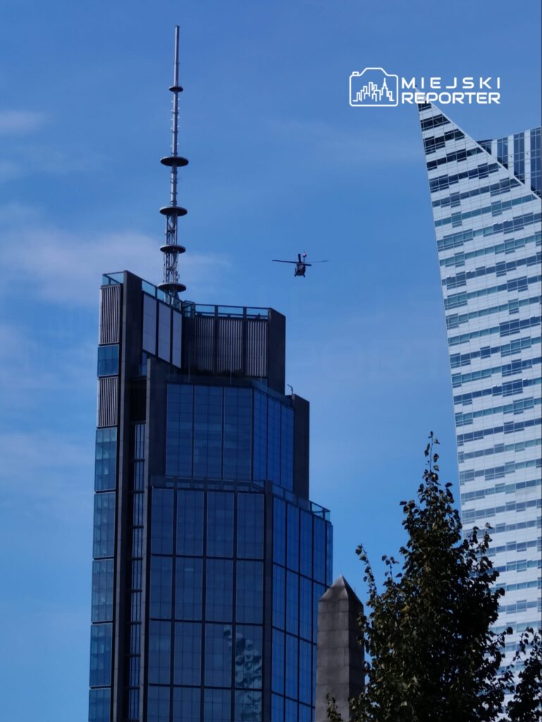 Helikopter przelatujący obok nowoczesnego biurowca z przeszkloną fasadą na tle błękitnego nieba.