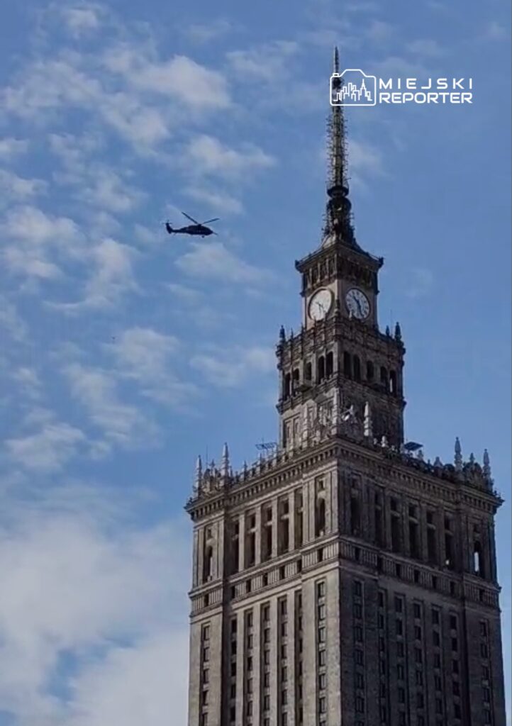 Helikopter przelatujący nad wieżowcem z zegarem w słoneczny dzień, z niebieskim niebem w tle.