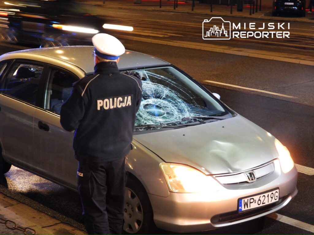 Policjant w mundurze bada uszkodzony samochód osobowy z wybitym przednim szkłem na ulicy nocą.