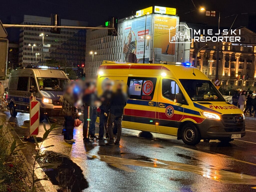 Grupa ludzi rozmawia z policjantem obok karetki pogotowia na ruchliwej ulicy w nocy.