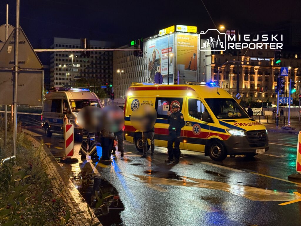 Na oświetlonej ulicy nocą, karetka pogotowia oraz radiowóz policji zatrzymały się przy grupie osób, z których jedna otrzymuje pomoc.