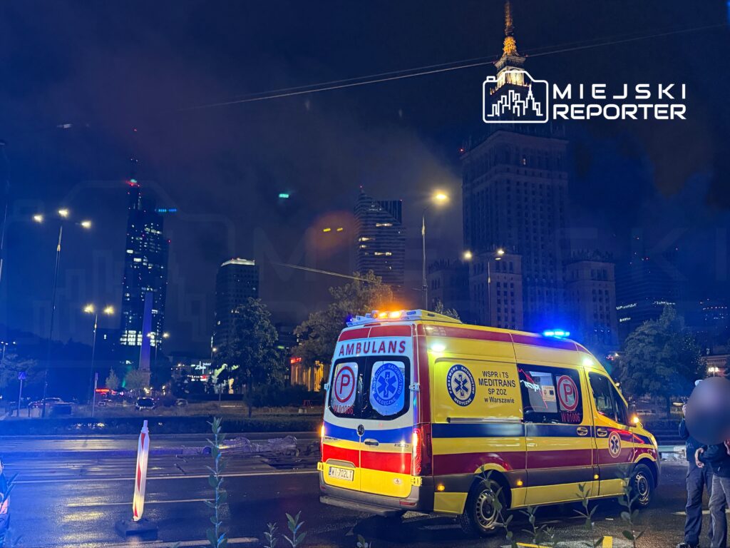 Karetka pogotowia z włączonymi sygnałami świetlnymi stoi na ulicy nocą, w tle widoczne nowoczesne wieżowce.