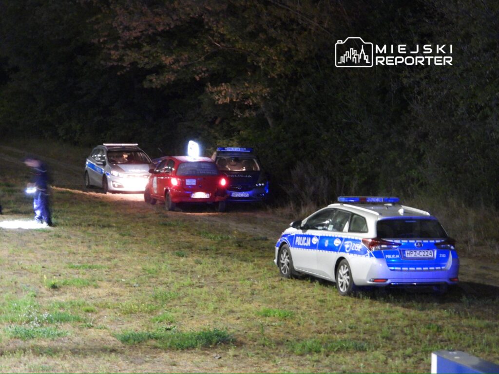 Policjanci w mundurach przeszukują teren przy zaparkowanych radiowozach w lesie nocą.