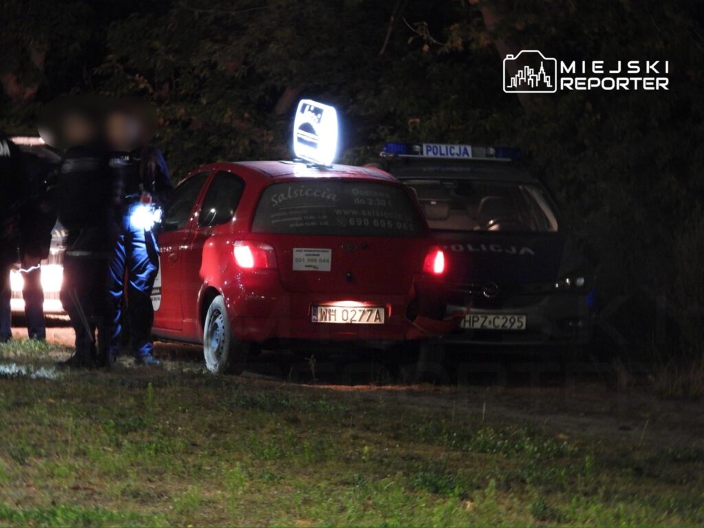 Grupa policjantów sprawdzająca czerwoną taksówkę na poboczu drogi w nocy, z włączonymi sygnałami świetlnymi.