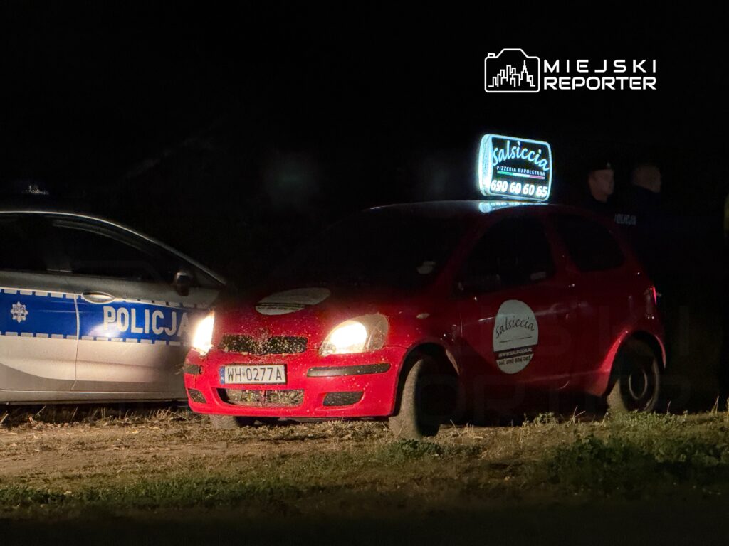 Czerwony samochód dostawczy z podświetloną reklamą pizzerii stoi obok radiowozu policji w ciemnym otoczeniu.
