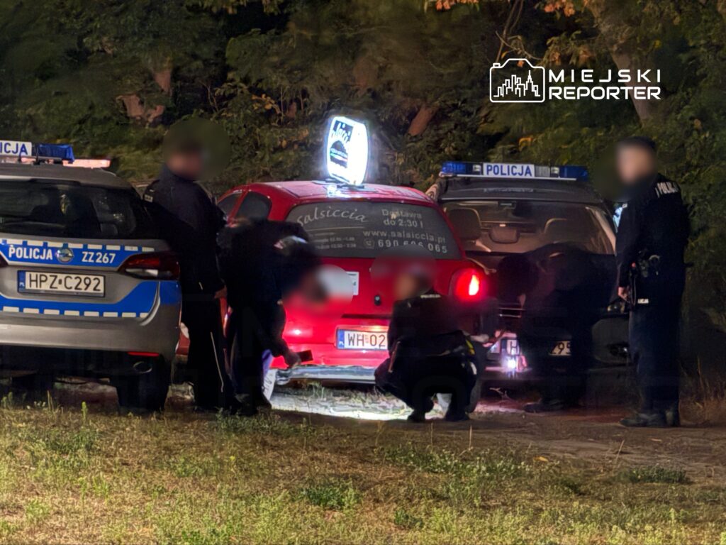 Grupa policjantów przeszukuje czerwoną furgonetkę zaparkowaną w ciemnym otoczeniu, obok dwóch radiowozów.
