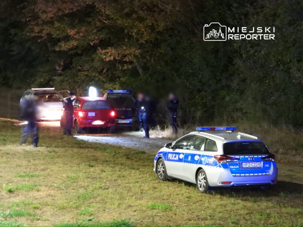 Policjanci prowadzą czynności dochodzeniowe przy zaparkowanych samochodach w lesistym terenie nocą.