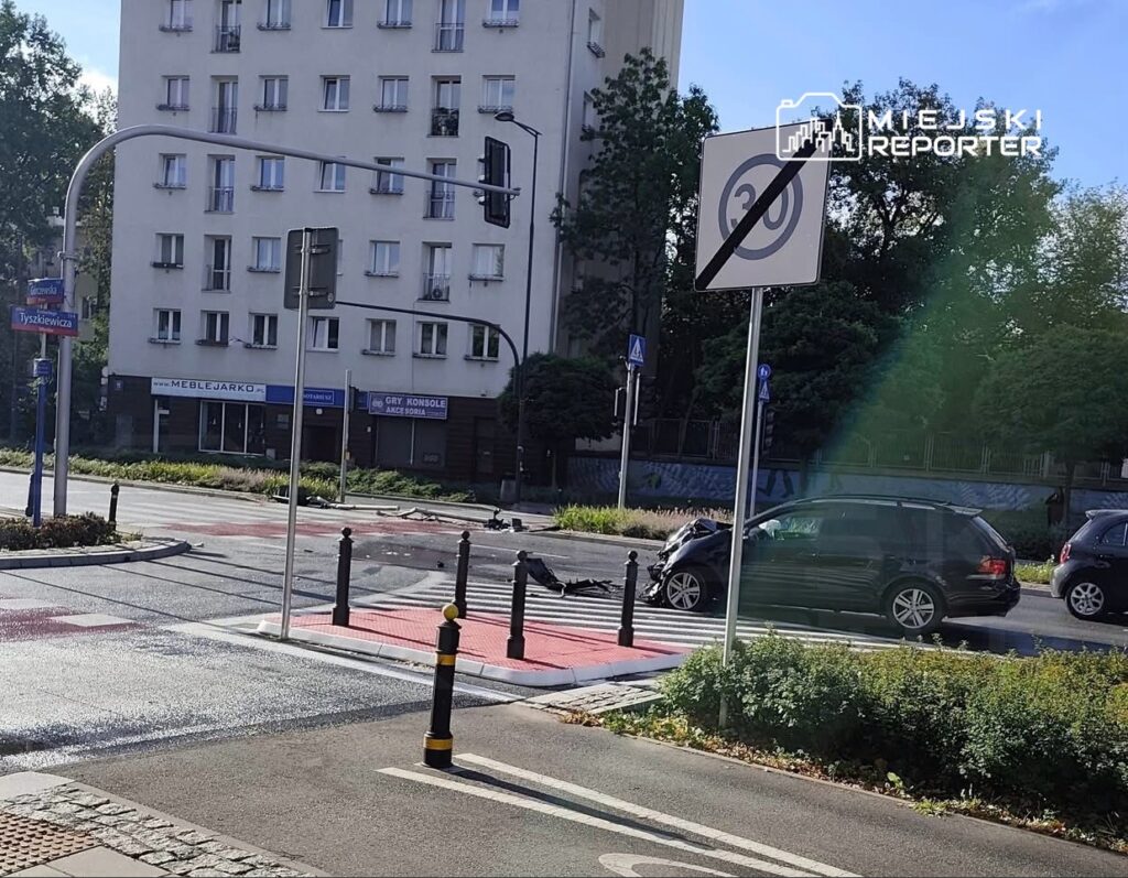 Zniszczony samochód na skrzyżowaniu, obok znak drogowy z ograniczeniem prędkości i budynek mieszkalny w tle.