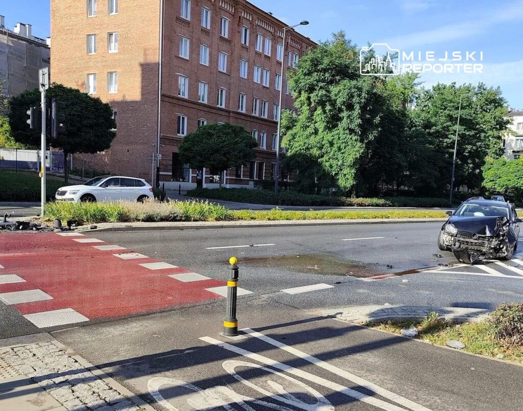 Zniszczony pojazd osobowy leży na skrzyżowaniu, obok nowoczesnego budynku mieszkalnego, w otoczeniu zieleni.
