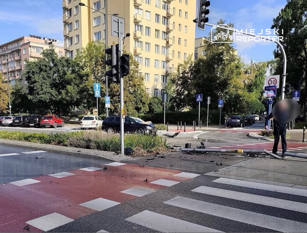 Na skrzyżowaniu zniszczony słup sygnalizacji świetlnej, a obok stoi mężczyzna w czarnym ubraniu, oceniając sytuację.