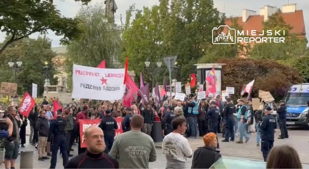 Grupa protestujących z transparentami i flagami, stojąca na ulicy w otoczeniu policji oraz pomnika.