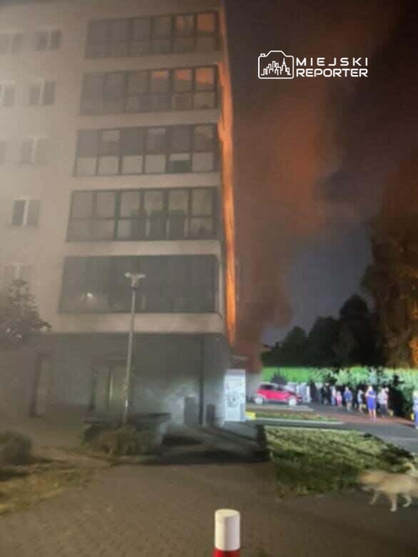 Płomienie wydobywające się z okna nowoczesnego budynku, obok grupy ludzi obserwujących sytuację.