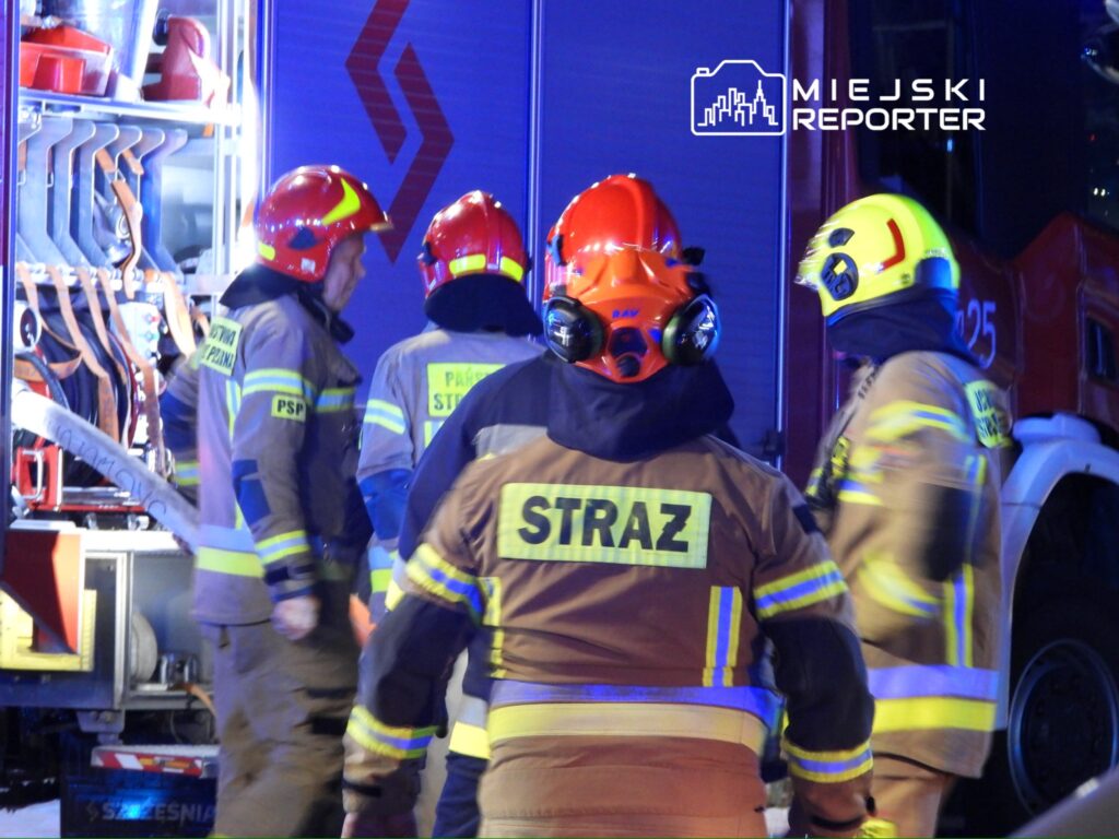 Grupa strażaków w mundurach z odblaskowymi pasami omawia działania przy wozie strażackim w nocy.