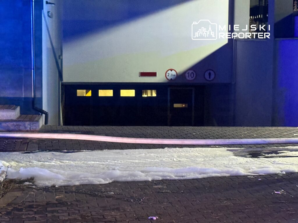 Biała piana pokrywająca chodnik przed garażem, z widocznymi znakami drogowymi i podświetleniem nocnym.
