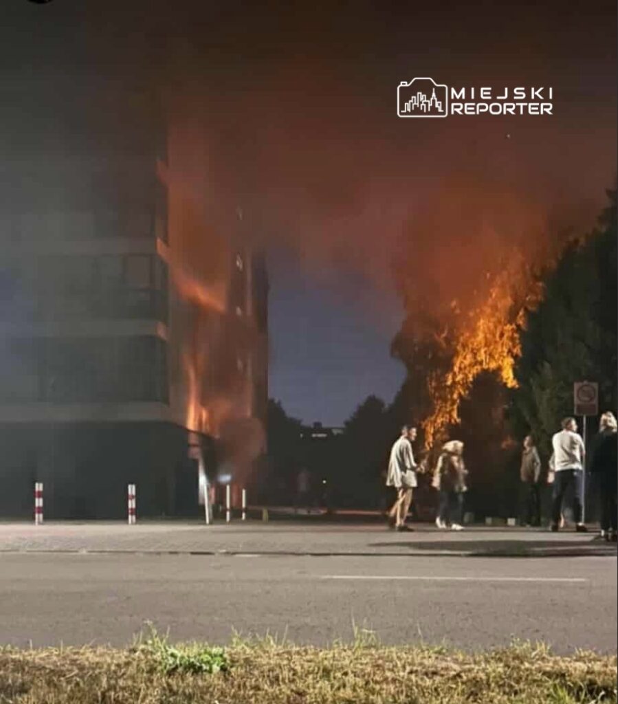 Ogień wydobywający się z budynku mieszkalnego, w tle grupa ludzi obserwująca sytuację na ulicy.