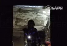 Strażak z latarką przeszukuje ciemne wnętrze budynku, obok niego inny ratownik w hełmie.