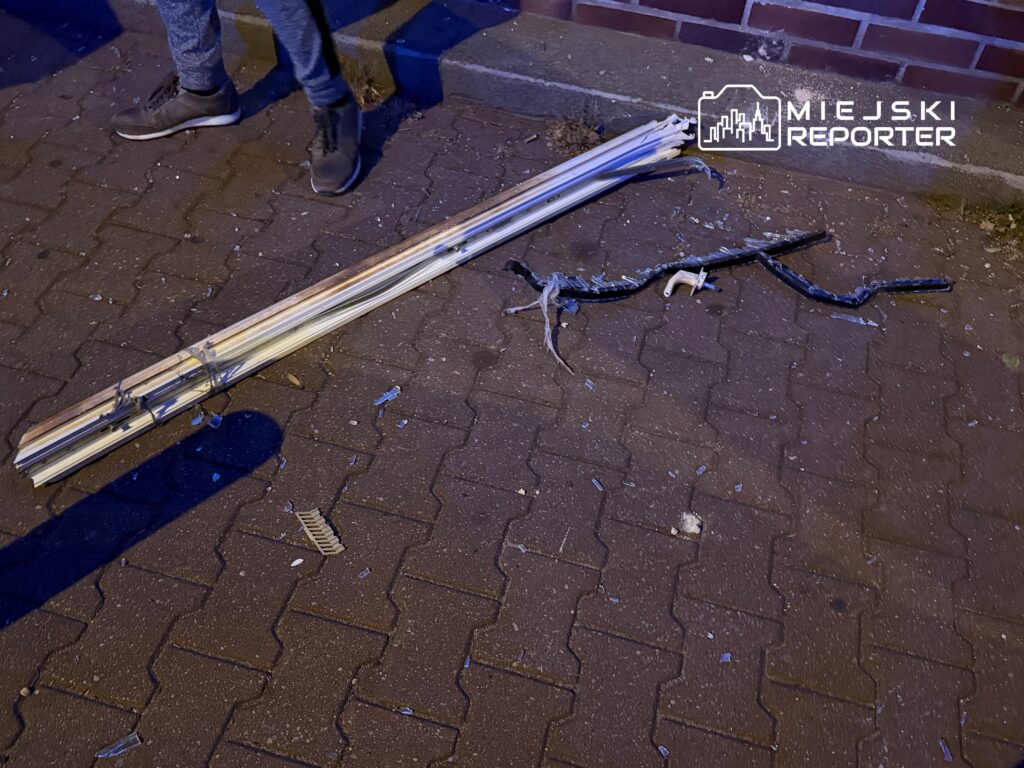 Na chodniku leżą kawałki rur i przewodów, obok nich fragmenty rozbitego szkła. W tle widoczna jest ceglana ściana budynku.
