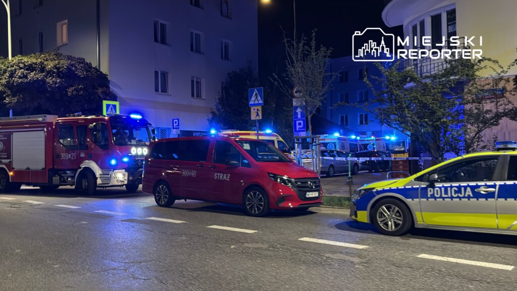 Na ulicy zaparkowane pojazdy straży pożarnej i policji, w tle widać migające niebieskie światła.