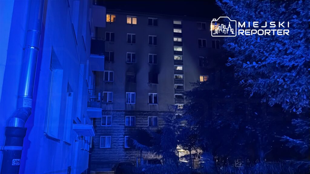 Widok na blok mieszkalny oświetlony niebieskim światłem w nocy, z otwartymi oknami na wyższych piętrach.