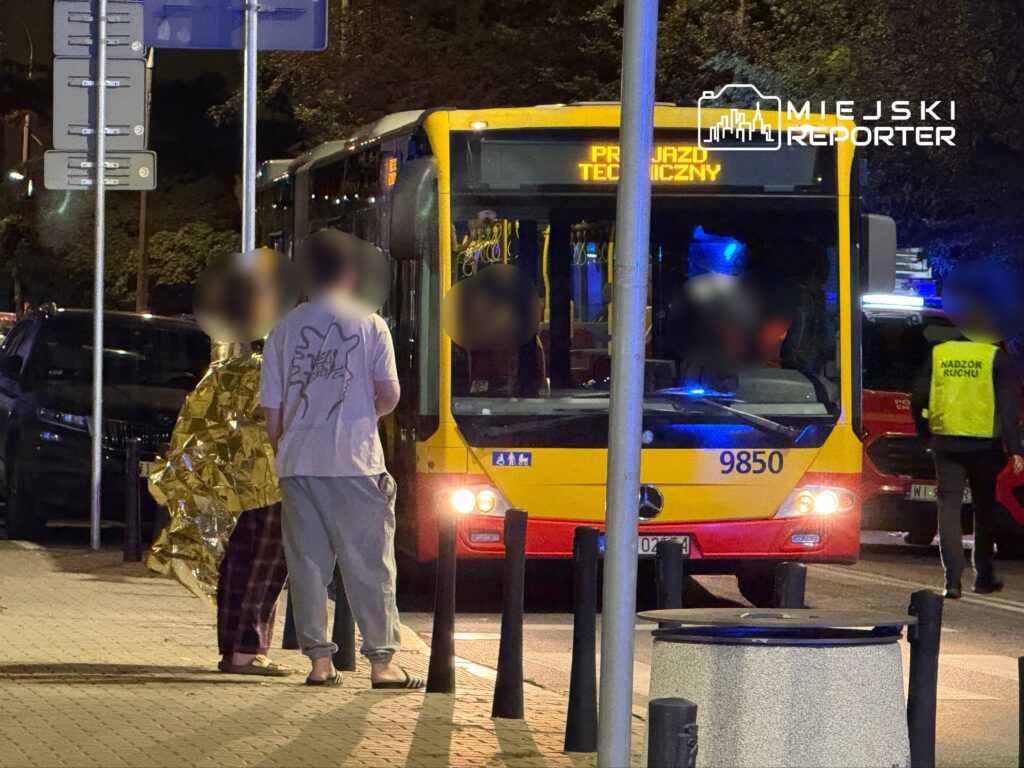 Dwójka ludzi w kocach ratunkowych rozmawia obok autobusu z napisem "PRZYJAŹŃ TECHNICZNY" na przystanku nocą.