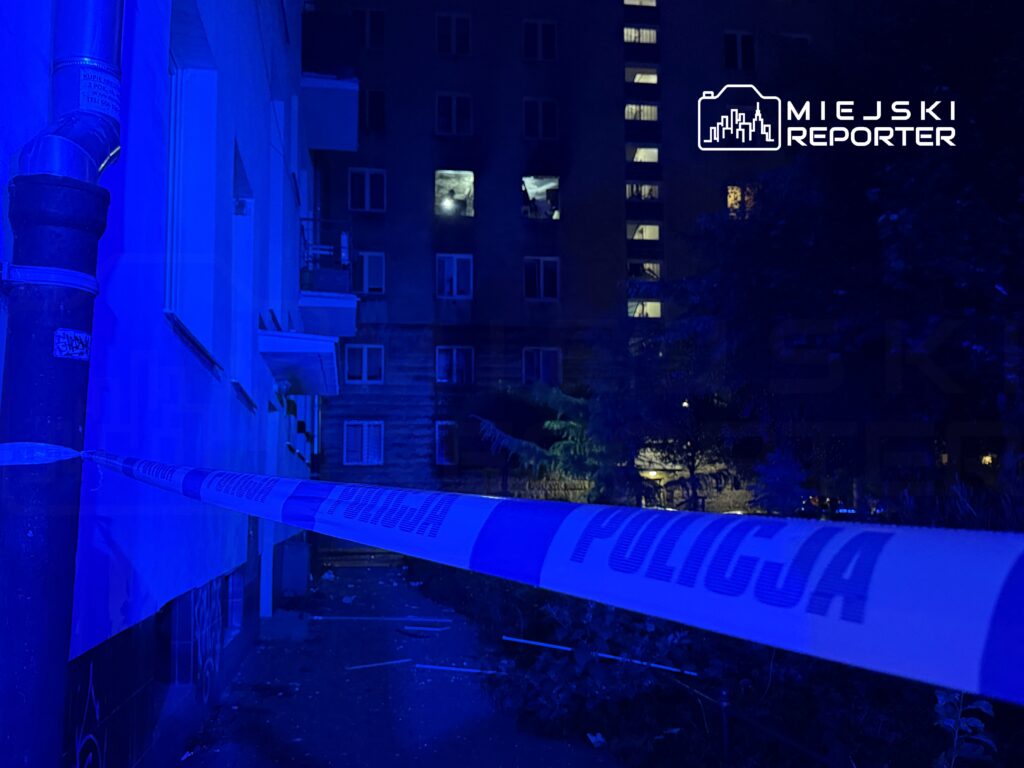 Nieoznakowane miejsce z niebieskim światłem, otoczone taśmą policyjną, z widocznymi oknami budynku w tle.