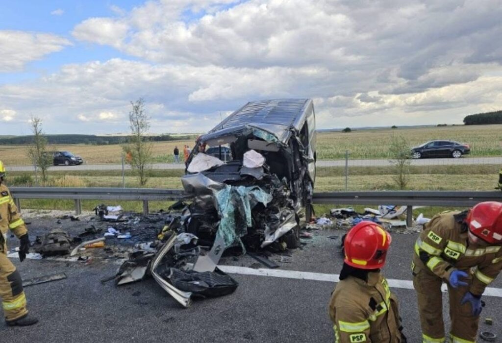 Strażacy w mundurach pracują przy zniszczonym pojeździe na drodze, obok leżą wraki i rozbite szkło.
