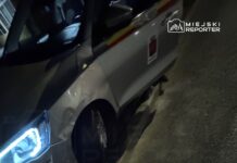 Samochód służbowy z uszkodzoną oponą stoi na poboczu drogi w nocy, w otoczeniu asfaltu i trawy.