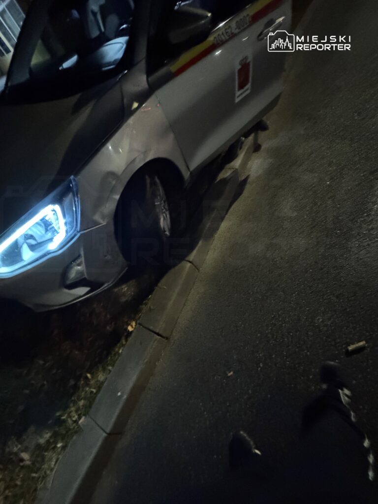 Samochód służbowy z uszkodzoną oponą stoi na poboczu drogi w nocy, w otoczeniu asfaltu i trawy.