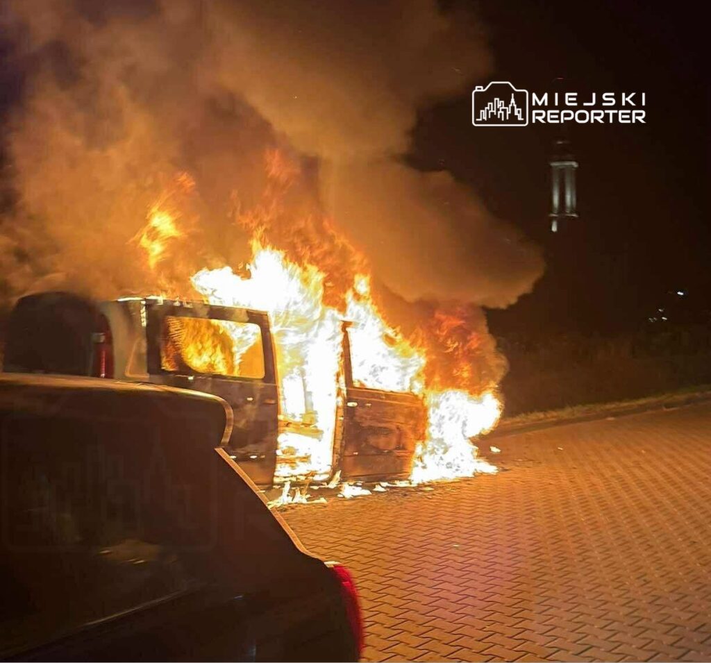 Pojazd w ogniu na parkingu, z płomieniami wydobywającymi się z wnętrza i gęstym dymem unoszącym się w powietrzu.