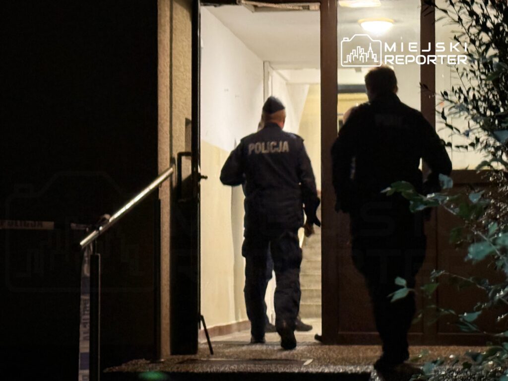 Dwóch policjantów w mundurach wchodzi do budynku przez otwarte drzwi w nocy.