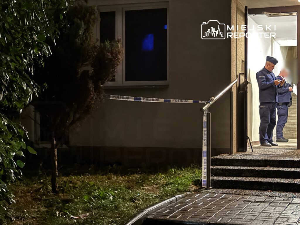 Dwóch policjantów stojących przy wejściu do budynku, obok taśmy zabezpieczającej. Nocna sceneria z roślinnością w tle.