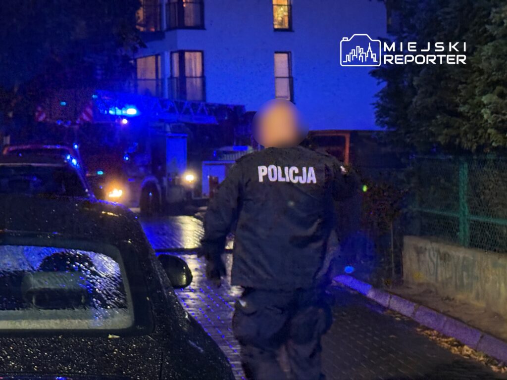 Policjant w mundurze z napisem "POLICJA" przechodzi obok zaparkowanych samochodów w oświetlonym niebieskim świetle.