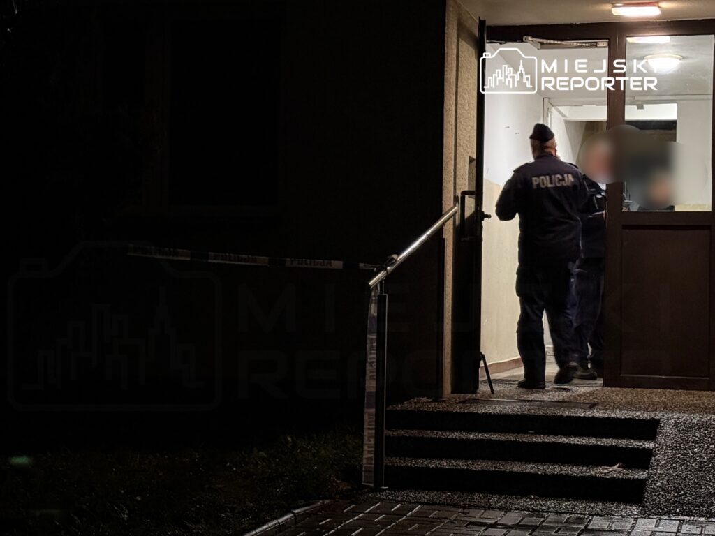 Dwaj policjanci w mundurach wchodzą do budynku przez otwarte drzwi, przy których znajduje się taśma zabezpieczająca.
