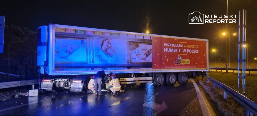 Trzech ratowników pracuje przy uszkodzonej ciężarówce na drodze w nocy, obok widać reklamę pieluszek.