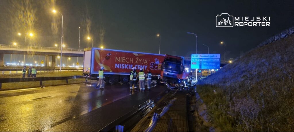 Ciężarówka przewrócona na autostradzie, w pobliżu grupy ratowników oraz oznakowania kierunkowego w nocy.