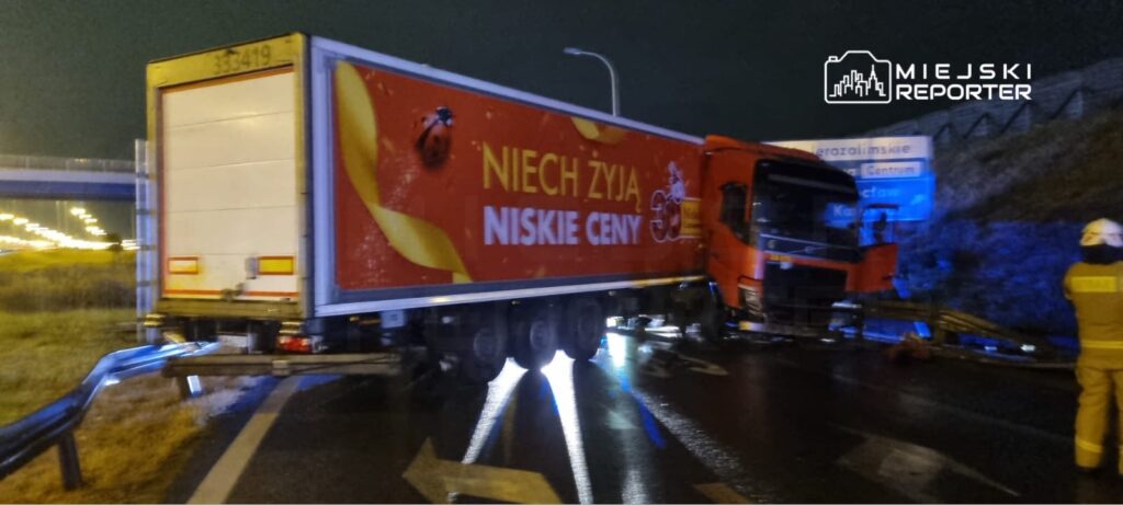 Czerwony ciężarówka z reklamą "Niech żyją niskie ceny" stoi na drodze po kolizji, obok widoczni strażacy.