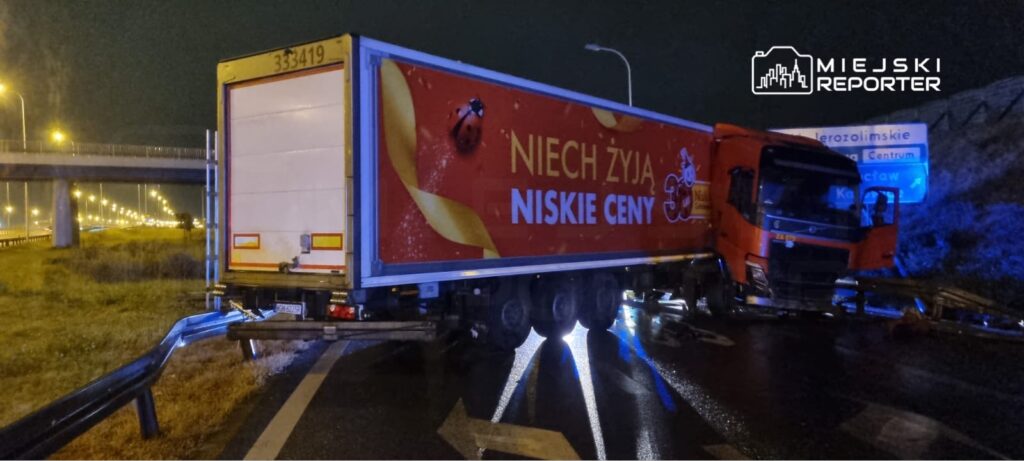 Czerwony ciężarówka z reklamą "NIECH ŻYJĄ NISKIE CENY" stoi na drodze, przy oświetlonej autostradzie, w nocy.