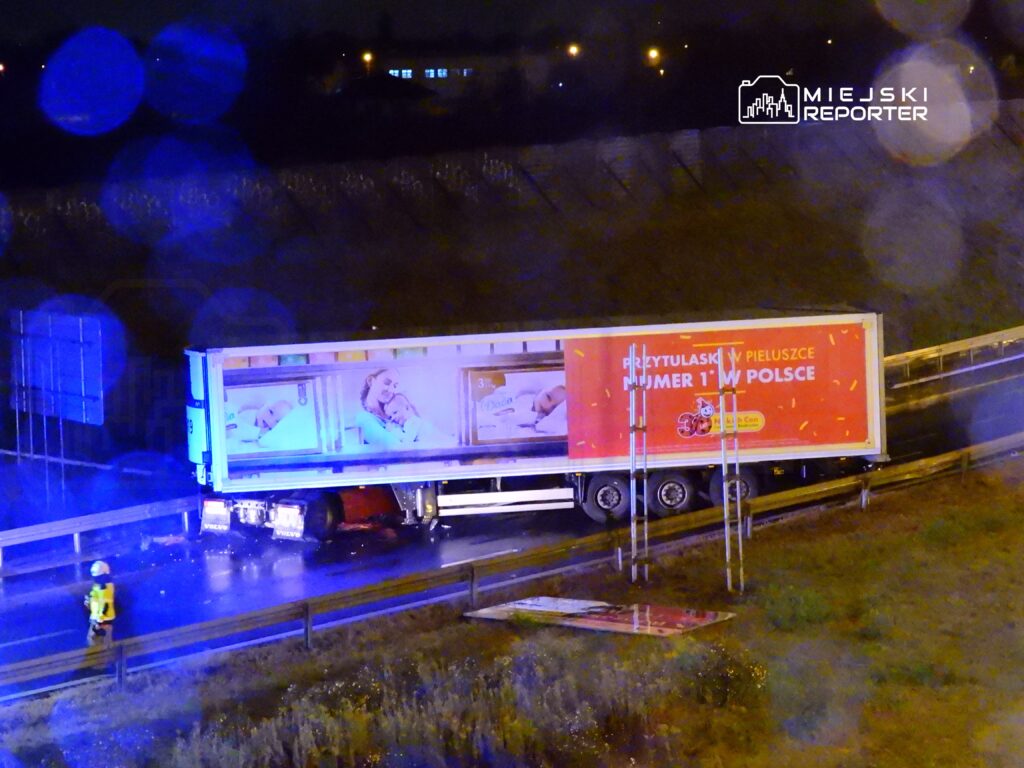 Czerwony ciężarówka na poboczu drogi, z reklamą pieluszek, obok pracujący strażak w kamizelce odblaskowej.